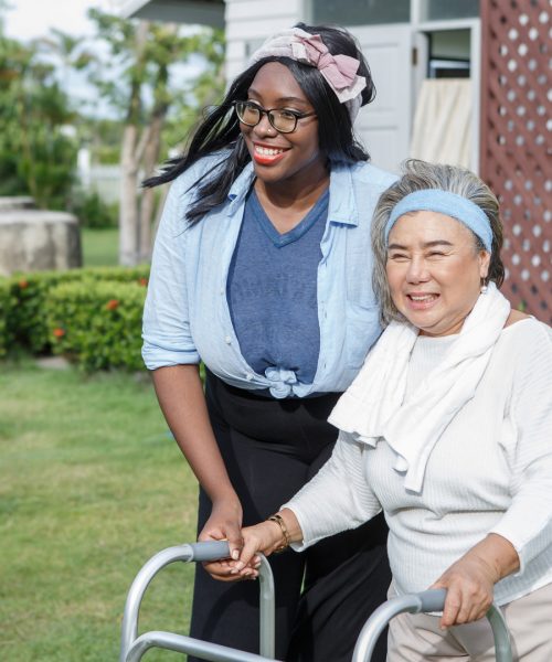 asian senior mother walk with walker and african american carer support in garden at home. old woman disabled walking and black caregiver young woman helping in back yard park outdoors. health care