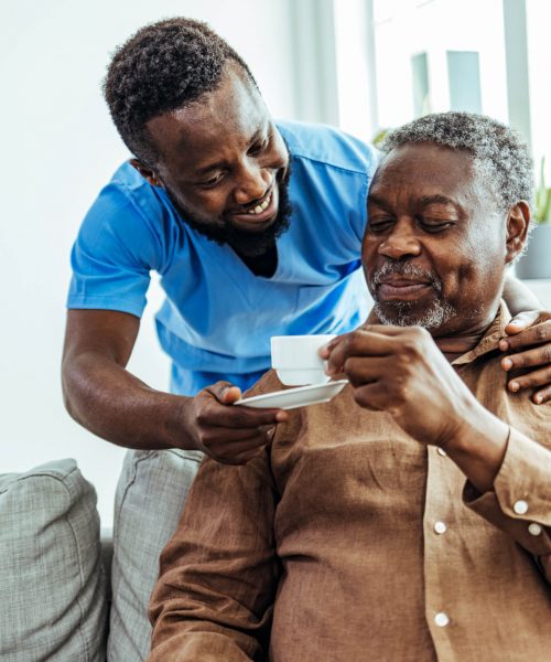 Home caregiver helping a senior man drinking a cup of coffee at a nursing home.